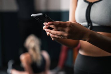 Bir kadın spor salonunda bir iletişim cihazı kullanıyor, elinde tutuyor, kolu açık ve başparmağı ekranda sallanıyor, bileği ve dirseği hafifçe bükük.