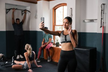 Bir kadın spor salonunda telefonuyla selfie çekiyor, kaslı kaslarını ve atletik fiziğini gösteriyor. Şort giyiyor ve eli uzatılmış, kolunu ve bacak kaslarını esnetiyor.