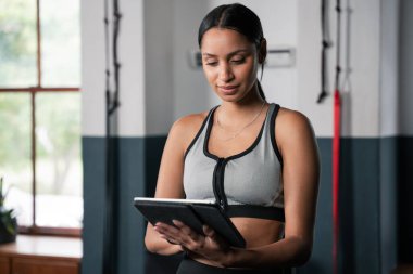 Sarışın bir kadın spor salonunda egzersiz yaparken kalçasında tablet alet kullanıyor. Yanında bir saksı çiçeği oturuyor ve arka planda bir kapı var.