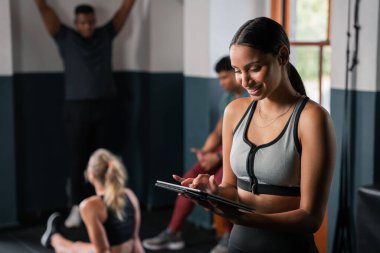 Bir kadın spor salonunda kol kaslarını esnerken rahatça bir tablet kullanıyor. Egzersiz videolarını incelerken eliyle jest yapıyor, diğer jimnastikçilerle eğlenceli egzersiz fikirleri paylaşıyor.