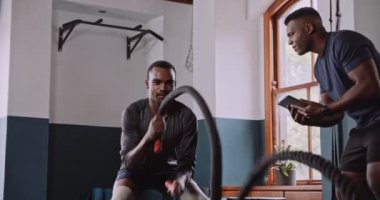 Spor salonundaki iki adam yoğun antrenman için halat kullanıyor, eklem güçlerini ve takım çalışmalarını sergiliyorlar. Odaklandıkları konu kas güçleri ve güçlü topluluk ruhları.