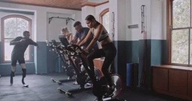 Bir grup birey spor salonunda egzersiz bisikletleri kullanırken ritmik davul sesleri odayı dolduruyor ve enerjik ve sanatsal bir atmosfer yaratıyor.