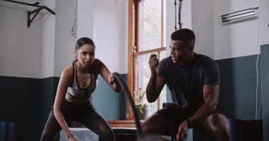 Bir adam ve bir kadın, arka planda çalan ahşap sandalyeler ve müzikle çevrili büyük bir pencerenin yakınındaki bir spor salonunda iple çalışıyor.