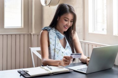 Kredi kartı tutan Asyalı genç bir kadın ve ev ofisteki internet ürünleri için dizüstü bilgisayar kullanıyor. Çevrimiçi alışveriş kavramı.