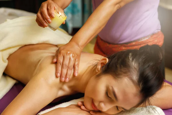 Masseuse drips warm massage oil from a candle on the back of the young woman.