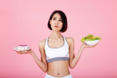 Genç bir kadın pembe arka planda izole edilmiş bir pasta ve sağlıklı salata arasında seçim yapıyor. Sağlıklı beslenme, beslenme, kilo verme konsepti.