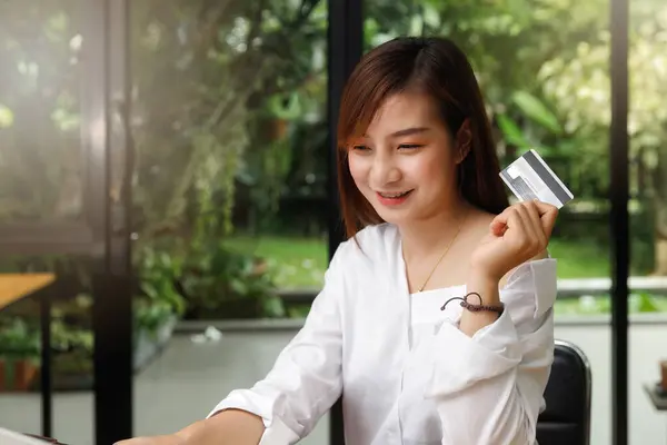 Smiling beautiful Asian woman presenting credit card in hand showing trust and confidence for making payment. Online shopping concept.