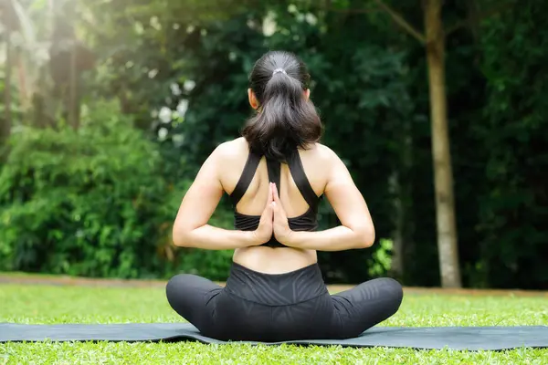 Asian woman practicing yoga in reverse prayer pose, back and shoulders stretching in outdoor park.