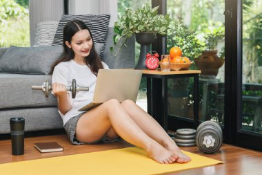 Dambılı güzel genç bir kadın oturma odasında otururken dizüstü bilgisayarından fitness videoları izliyor..