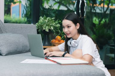 Güzel Asyalı kadın gülümsüyor ve oturma odasında dizüstü bilgisayara bakıyor. Çalışmak, ev kavramından öğrenmek.