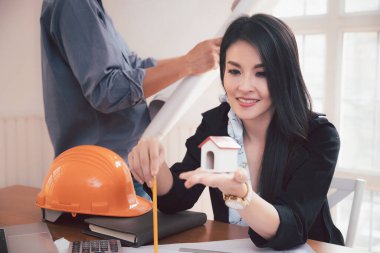 Bir kadın iş yerinin önünde otururken ev modelini tutuyor. Ofiste planla çalışan iki iş arkadaşından oluşan bir grup. Eş zamanlı takım çalışması kavramı.