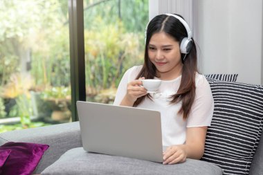Pretty smiling young woman wear headphones sitting on sofa holding a cup of coffee and usinging on tablet.
