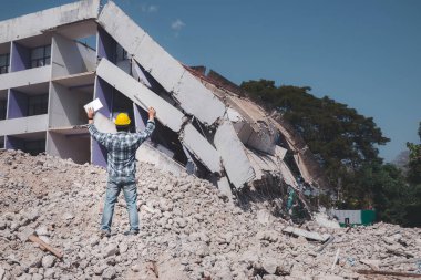 Tablet ve mavi baskıyla ayakta duran bir mühendis yıkılan binanın yıkıntıları üzerine elini kaldırıyor..