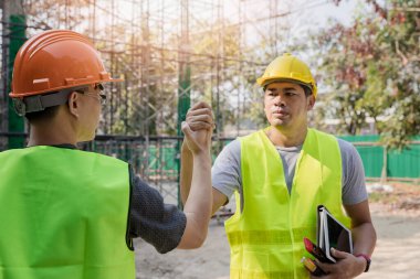 İnşaatçılar inşaat alanında el sıkışarak birbirlerini selamlıyorlar. Koruyucu kasklı ve yelekli inşaat işçileri el ele tutuşuyorlar. Takım çalışması, ortaklık, jest konsepti.