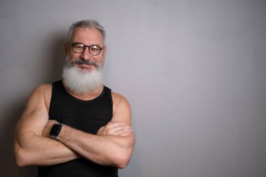 Siyah kolsuz tişörtlü, beyaz sakallı, kolları göğsünde, gri bir arka planda çapraz duran yaşlı bir adam. Her yaştaki fiziksel ve fiziksel aktiviteleri vurgular.