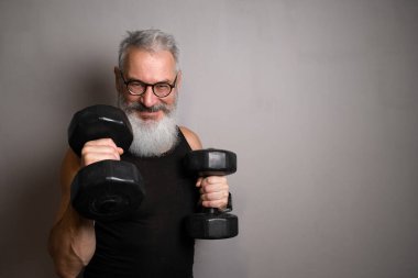 Siyah askılı beyaz sakallı yaşlı bir adam, gri arka planda halterleri tutuyor. Her yaştaki aktif yaşlanma ve sağlığı simgeler..