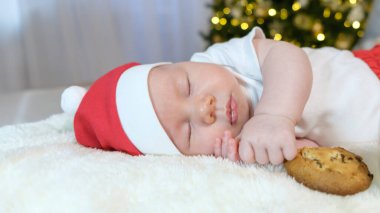 İki aylık bir bebek, şenlikli bir Noel kıyafeti giymiş, parlak bir arka planda süt ve kurabiyelerle uzanmış, bayram neşesi ve konforu yayıyor..