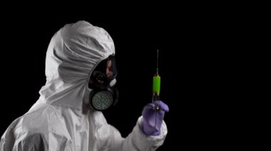Kimyasal koruma giysisi içinde bir laboratuvar çalışanı elinde siyah bir arka plan üzerinde yeşil enjeksiyonlu bir şırınga tutuyor.