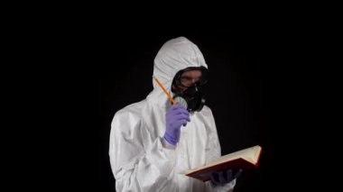 Kimyasal koruma giysisi içinde bir laboratuvar çalışanı siyah arka planda bir kalemle bir deftere yazıyor.