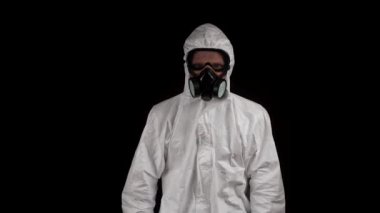 Kimyasal korumalı bir laboratuvar çalışanı siyah arka planda kameraya gider.