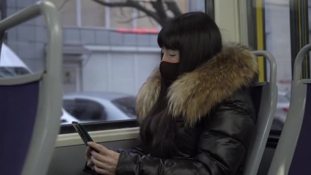 女の子はコロナウイルスの流行中にマスクを着用するトラムに乗ります スマートフォンを見ている女の子 スローモーション — ストック動画