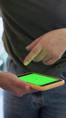 Elinde yeşil ekranlı bir akıllı telefon tutan bir kadın, sıradan bir düzende parmaklarıyla arayüzde geziniyor.