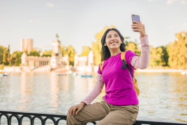 Emeklilik parkında gölün önünde akıllı telefonuyla selfie çeken gülümseyen turist, Madrid