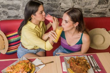 Two women sharing drinks and enjoying a traditional ecuadorian meal at a restaurant in Portoviejo, savoring the flavors of latin american cuisine clipart