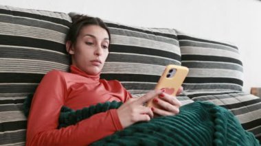Kanepede uzanmış genç bir kadın, akıllı telefonu yavaş çekimde karıştırıyor, rahatlamanın ve bağlanmanın tadını çıkarıyor.
