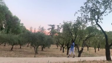 Spor kadın huzurlu parkta yürüyor, günbatımının tadını çıkarırken kablosuz kulaklıklar ve akıllı kulaklıkla müzik dinliyor.