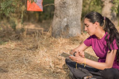 Genç Latin kadın oryantasyon yarışına katılıyor, zamanı kontrol ediyor ve ormandaki haritayı kontrol ediyor.