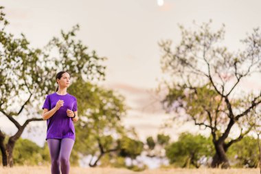 Latin bir kadın Madrid parkında koşuyor, doğa, ağaçlar ve günbatımının ılık parıltısıyla çevrili aktif bir yaşam tarzını kucaklıyor.