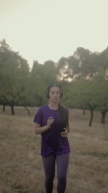 Genç Latin kadın müzik dinlerken altın saat boyunca manzaralı parkta koşuyor. Mor spor giysisiyle sakin ve motive edici bir ifade takınıyor.