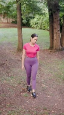 Güneşli parkta yürüyen ve esneyen atletik Latin kadın ağaçlar ve çimenlerle dolu yeşil bir arazide spor yapıyor.