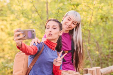 İki bayan yürüyüşçü ormanda akıllı bir telefonla selfie çekiyorlar, gülümsüyorlar ve barış işareti yapıyorlar.