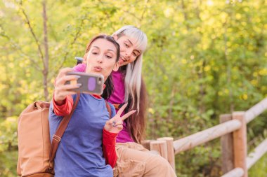 İki kadın yürüyüşçü, canlı bir ormanda yürürken akıllı telefonlarıyla mutlu bir şekilde selfie çekiyorlar ve doğadaki maceralarını yakalıyorlar.