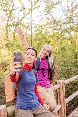 İki kadın yürüyüşçü, ormanda yürürken akıllı telefonlarıyla selfie çekiyorlar. Bir tanesi kameraya el sallıyor.