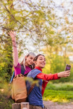 Sırt çantalı iki genç kadın ormanda yürüyüş yaparken akıllı telefonlarıyla selfie çekiyorlar ve kameraya el sallıyorlar.