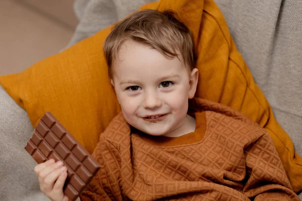 可爱的小男孩坐在家里的沙发上 吃着巧克力棒 孩子和糖果 糖糖果 孩子们享受美味的甜点 穿着休闲装的学龄前儿童 积极的情绪 — 图库照片