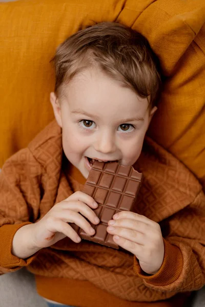可爱的小男孩坐在家里的沙发上 吃着巧克力棒 孩子和糖果 糖糖果 孩子们享受美味的甜点 穿着休闲装的学龄前儿童 积极的情绪 — 图库照片