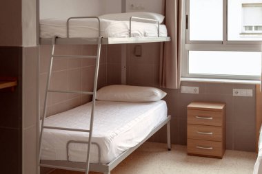 Temiz ve minimalist bir pansiyon odası ranzası, beyaz yatak, ve küçük ahşap bir komodin, uygun fiyatlı konaklama arayanlar için ideal. Ucuz pansiyonlar, tasarruf.