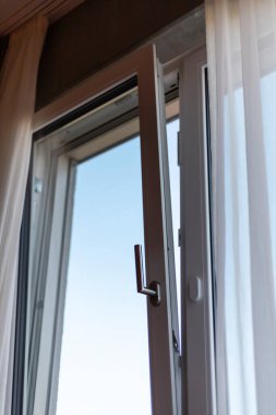 Beyaz çerçeveli pencere açık, güneş ışığının odayı aydınlatmasına olanak sağlıyor. Perdeler nazikçe sallanıyor, açık mavi bir gökyüzü ortaya çıkıyor. Sahne huzurlu bir gündüz atmosferini gösteriyor..