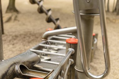 Metal bir çeşme oyun parkı ortamında su damlacıkları salıyor. Yakınlarda, ağaçlar ve yumuşak zemin ile çevrili oyun alanı ekipmanları sıcak güneşin altında parlıyor..