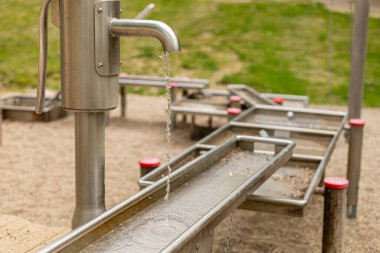 Paslanmaz çelik bir su pompası oyun parkındaki bir kanala su pompalar ve açık havada akan suyun eğlencesini keşfettiklerinde çocuklar için ilgi çekici bir deneyim yaratır..