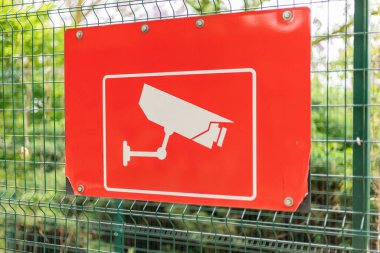 Beyaz mobese kamera grafikli parlak kırmızı bir tabela yemyeşil bir çitin ortasına asıldı. İşaret bölgedeki gözetleme tedbirleri hakkında bilgi veriyor..