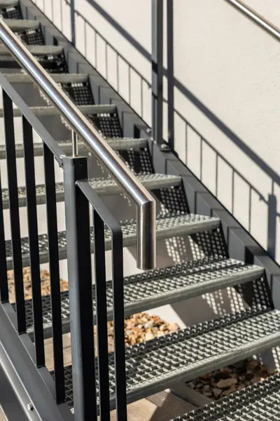 Sağlam tırabzanlar ve dokulu basamaklar içeren modern çelik merdivenlerin detaylı görüntüsü. Güneş ışığı tasarımı aydınlatır, kullanılan temiz çizgileri ve çağdaş materyalleri vurgular. Metal yapı