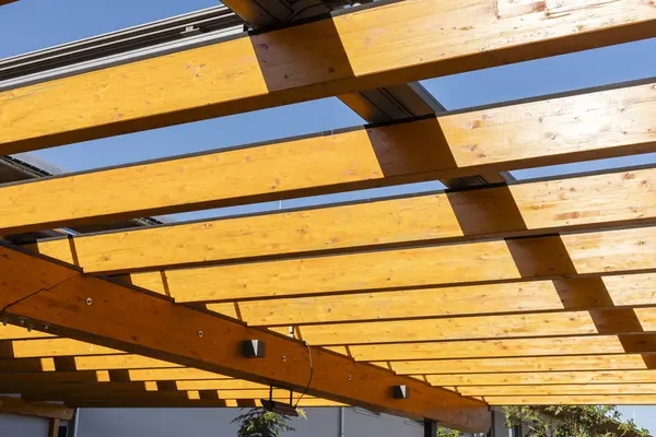 Tahta kirişler açık havada gölge sağlayan güneş koruma sistemini destekler. Açık mavi gökyüzü, güneşli bir günde yapıların tasarım ve işlevlerini vurgular..