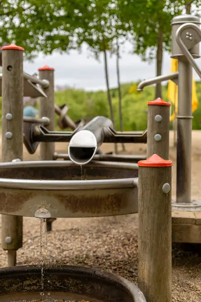 Çocuklar yaratıcılığı ateşlemek için tasarlanmış çeşitli etkileşimli özelliklerle donatılmış bir su oyun alanının keyfini çıkarırlar. Gökyüzü bulutlu bir öğleden sonra yeşilliklerle çevrilidir..