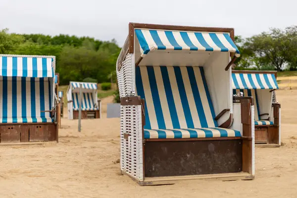 Mavi ve beyaz çizgili birkaç plaj sandalyesi kumlu bir plajda düzenlenir. Sahne bulutlu bir günde yeşilliklerle çevrili sessiz bir anı yakalıyor..