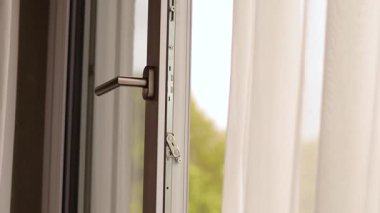 Güneşli bir odada açık bir pencere, temiz havanın içeri akmasına olanak sağlarken, yumuşak perdeler rüzgarla hafifçe hareket ederek huzurlu bir atmosfer yaratıyor..